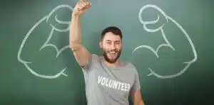 Man cheering while wearing a shirt reading "VOLUNTEER" and standing in front of a chalk board.