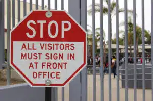 Sign on fence reading , "Stop. All visitors must sign in at front office".